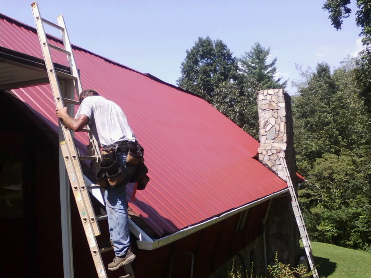 Roofer on a ladder beside a red metal roof and stone chimney — Soffit & Fascia Repair by Roofing Brothers in Duquesne